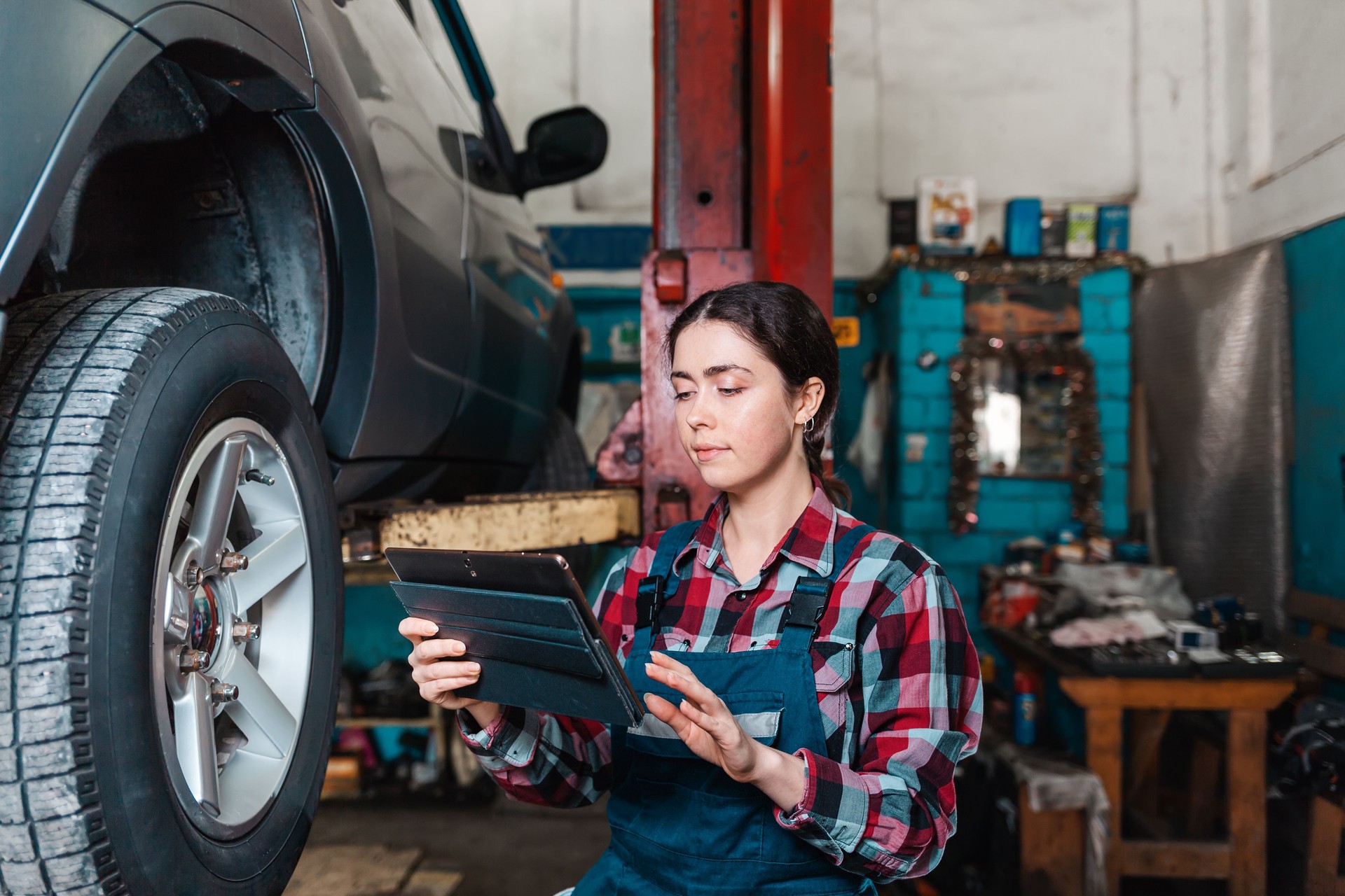 Portrait of a young female mechanic in uniform posing with a tablet in her hands. In the background is an auto repair shop. The car on the lift to the right